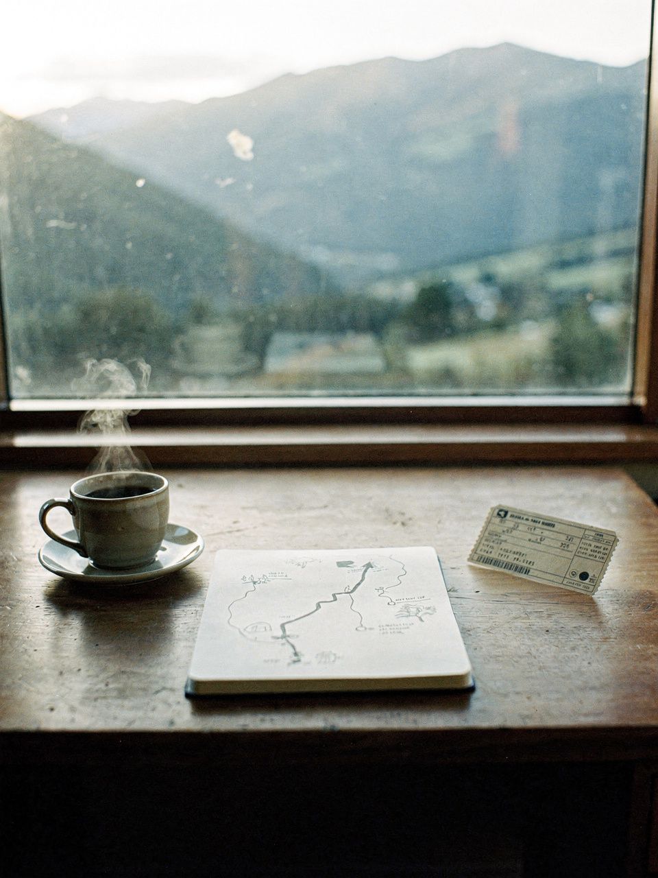 A desk by a window in Nelson: an open notebook with a hand-drawn route, a coffee, a paper ticket, the Tasman hills through the glass