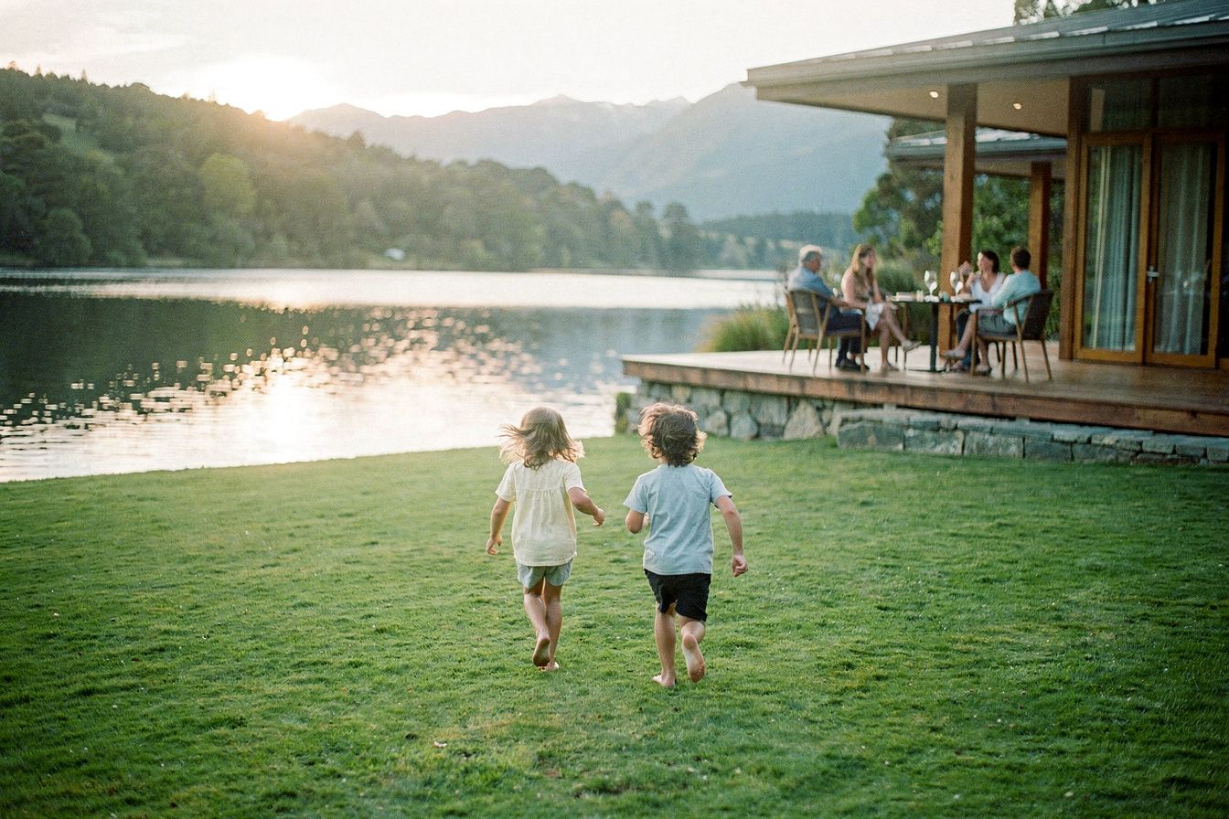 Two children running across a lawn towards a calm lake, adults on a lodge terrace behind