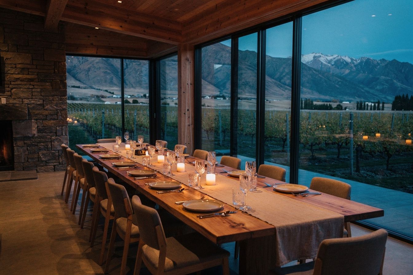 A lodge dining room in Central Otago at dusk, candlelight, windows onto vineyard rows and mountains