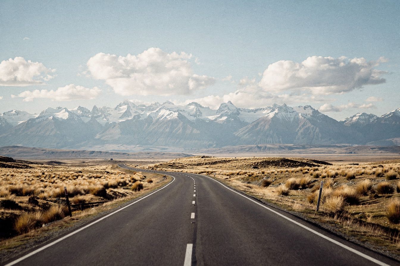 An empty two-lane road through golden tussock towards snow-capped mountains in the Mackenzie Basin